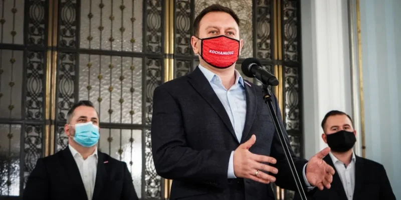 Trzech mężczyzn w garniturach stoi przy mikrofonie podczas konferencji prasowej, a centralny mężczyzna przemawia, pozostali stoją z boku, wszyscy w maseczkach.