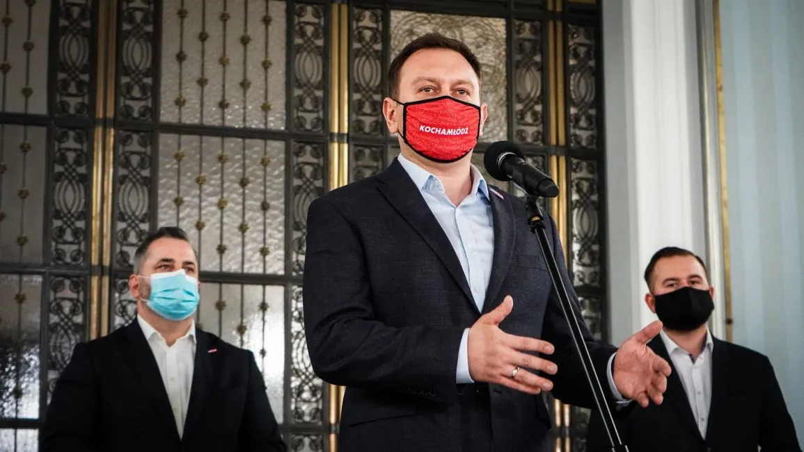 Trzech mężczyzn w garniturach stoi przy mikrofonie podczas konferencji prasowej, a centralny mężczyzna przemawia, pozostali stoją z boku, wszyscy w maseczkach.