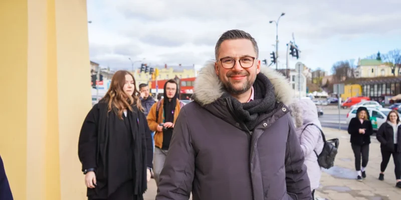 Na zdjęciu widać mężczyznę w ciepłej kurtce i okularach, który uśmiecha się, idąc w towarzystwie innych osób, na tle miejskiego krajobrazu.