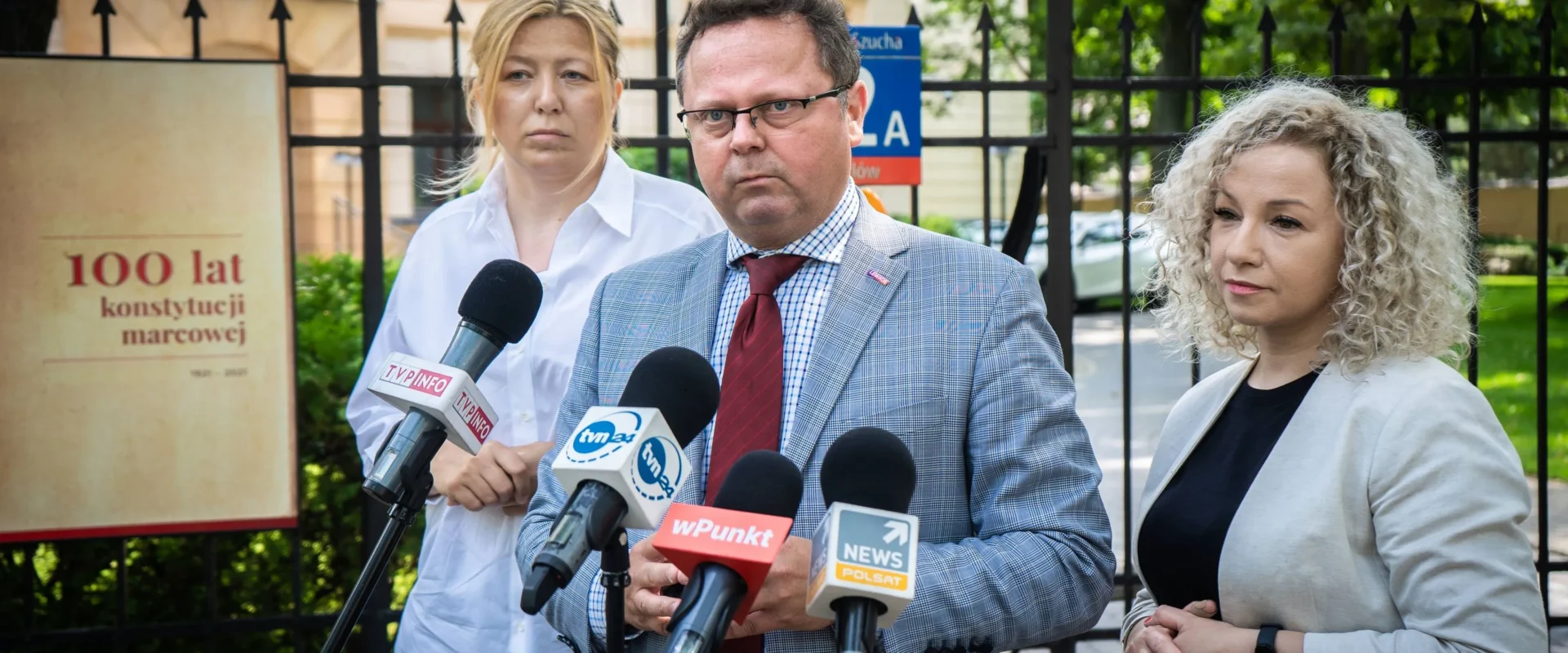 Trzy osoby stojące na zewnątrz budynku podczas konferencji prasowej, mężczyzna w garniturze z czerwonym krawatem przemawia do mikrofonów, a po jego bokach stoją dwie kobiety.
