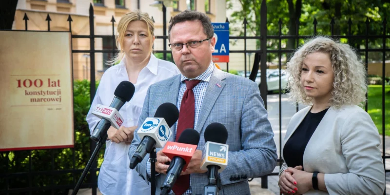 Trzy osoby stojące na zewnątrz budynku podczas konferencji prasowej, mężczyzna w garniturze z czerwonym krawatem przemawia do mikrofonów, a po jego bokach stoją dwie kobiety.