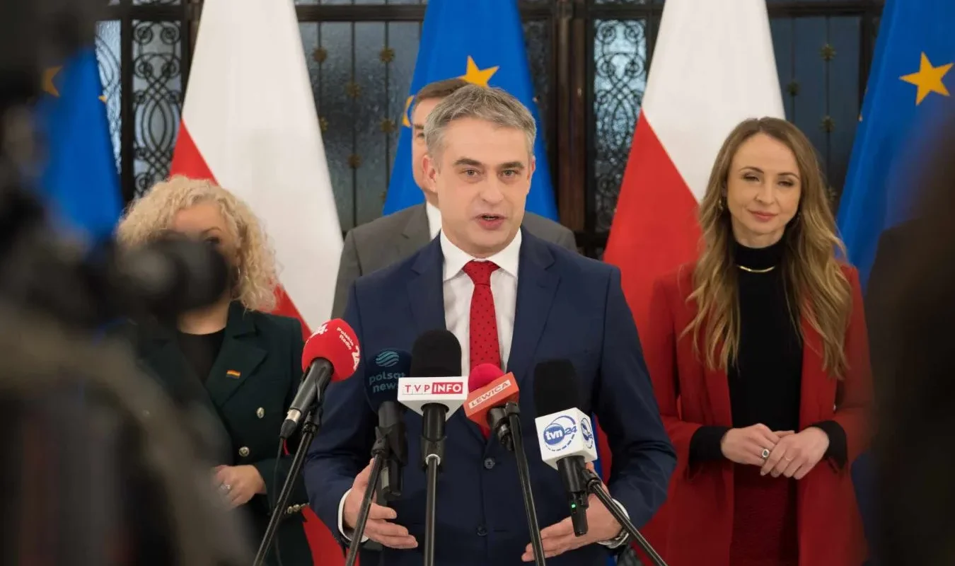 Polityk lewicy w granatowym garniturze przemawia na konferencji prasowej, stojąc za mikrofonami, z flagami Polski i Unii Europejskiej w tle.
