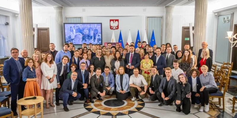 Duża grupa uczestników stojąca i siedząca w eleganckiej sali konferencyjnej z kolumnami, flagami UE i Polski oraz godłem Polski w tle, podczas podsumowania Okrągłych Stołów wokół zdrowia psychicznego dzieci i młodzieży w Polsce.
