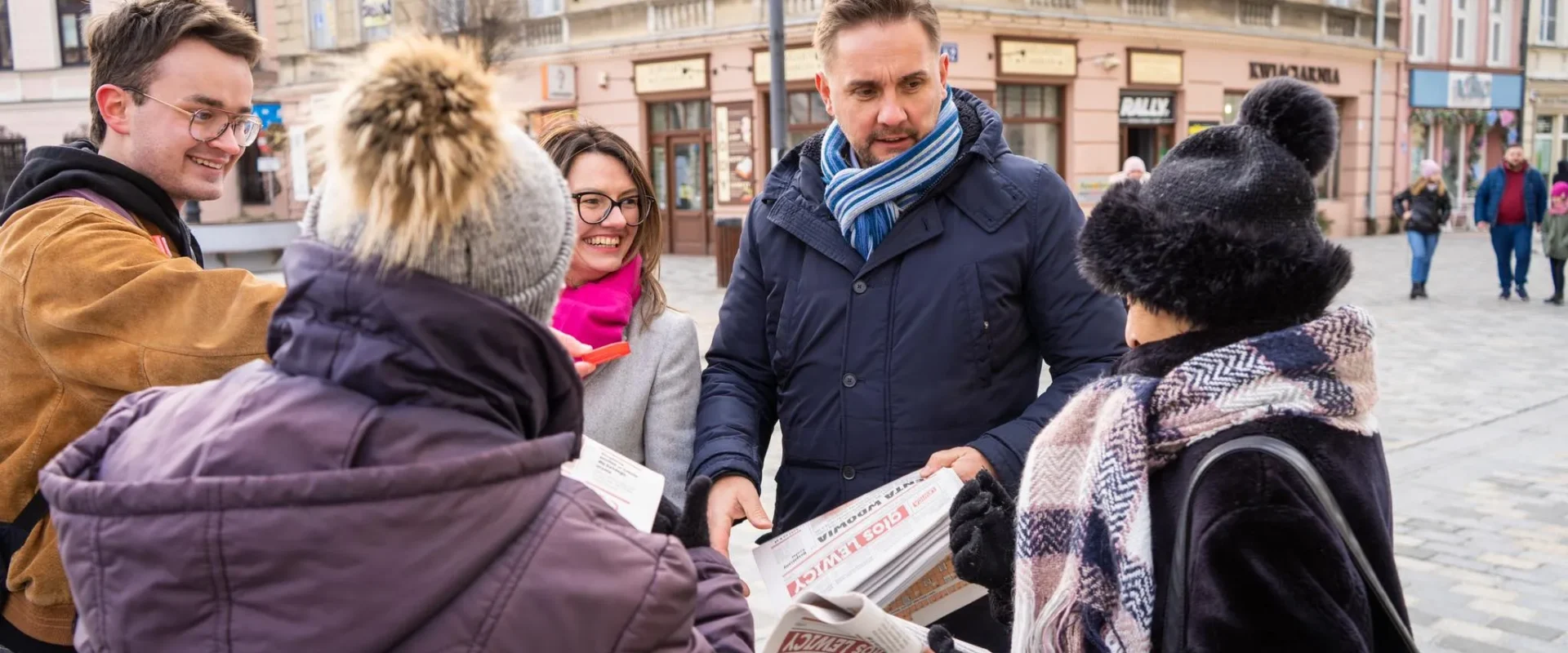 Grupa ludzi w zimowych ubraniach stoi na placu w mieście, rozmawia i rozdaje gazety wśród zabytkowych kamienic.