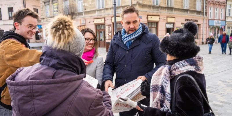 Grupa ludzi w zimowych ubraniach stoi na placu w mieście, rozmawia i rozdaje gazety wśród zabytkowych kamienic.