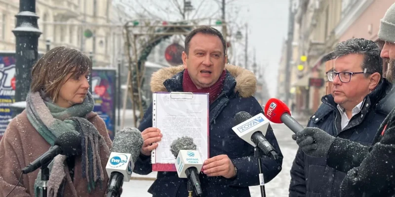 Mężczyzna w zimowej kurtce z futrzanym kapturem trzyma w dłoni dokument w teczce i zabiera głos do mikrofonów dziennikarzy na ulicy podczas zimowej konferencji prasowej.