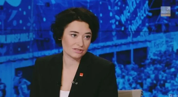 Kobieta w czarnej marynarce i białej bluzce siedzi w studiu telewizyjnym przed niebieskim, rozmytym tłem z wizerunkiem tłumu, wypowiadając się do kamery.