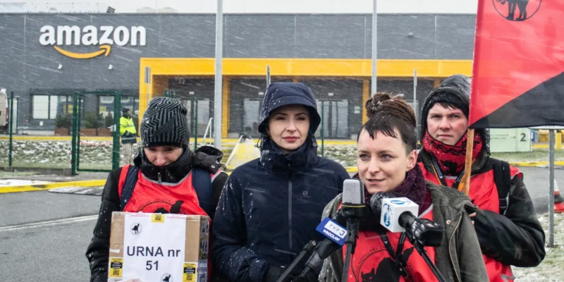 Grupa protestujących w czerwonych kamizelkach, z flagą, przemawia przed budynkiem Amazonu w śnieżnej scenerii.