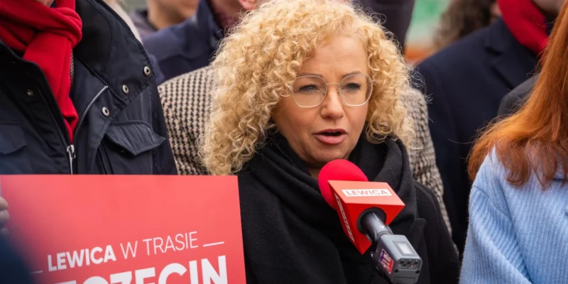 Kobieta z kręconymi blond włosami i okularami przemawia do mikrofonu z logo Lewica podczas demonstracji, stojąc obok czerwonego plakatu Lewica w Trasie.