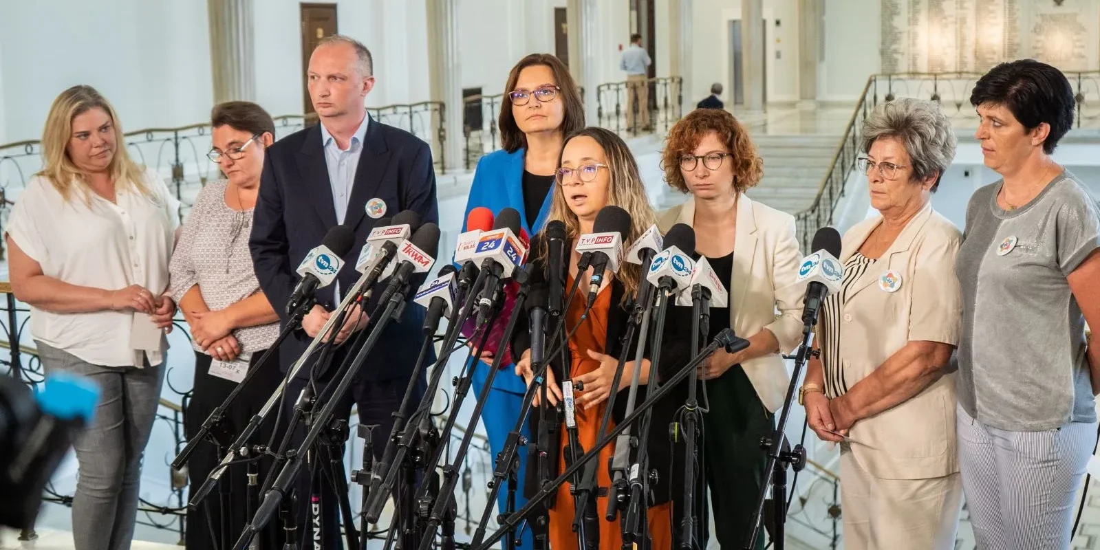 Grupa osób stoi na konferencji prasowej w holu, za licznymi mikrofonami, omawiając outsourcing w szpitalach.
