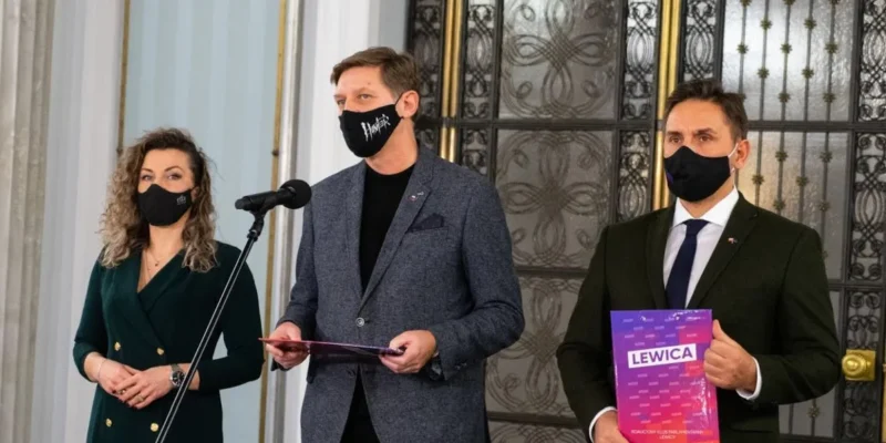 Trzy osoby w maseczkach stoją przed mikrofonem na konferencji prasowej: po lewej kobieta w zielonej sukni, na środku mężczyzna w szarym blazerze trzyma dokumenty, a po prawej mężczyzna w garniturze trzyma plik z napisem Lewica.