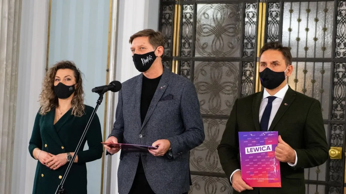 Trzy osoby w maseczkach stoją przed mikrofonem na konferencji prasowej: po lewej kobieta w zielonej sukni, na środku mężczyzna w szarym blazerze trzyma dokumenty, a po prawej mężczyzna w garniturze trzyma plik z napisem Lewica.