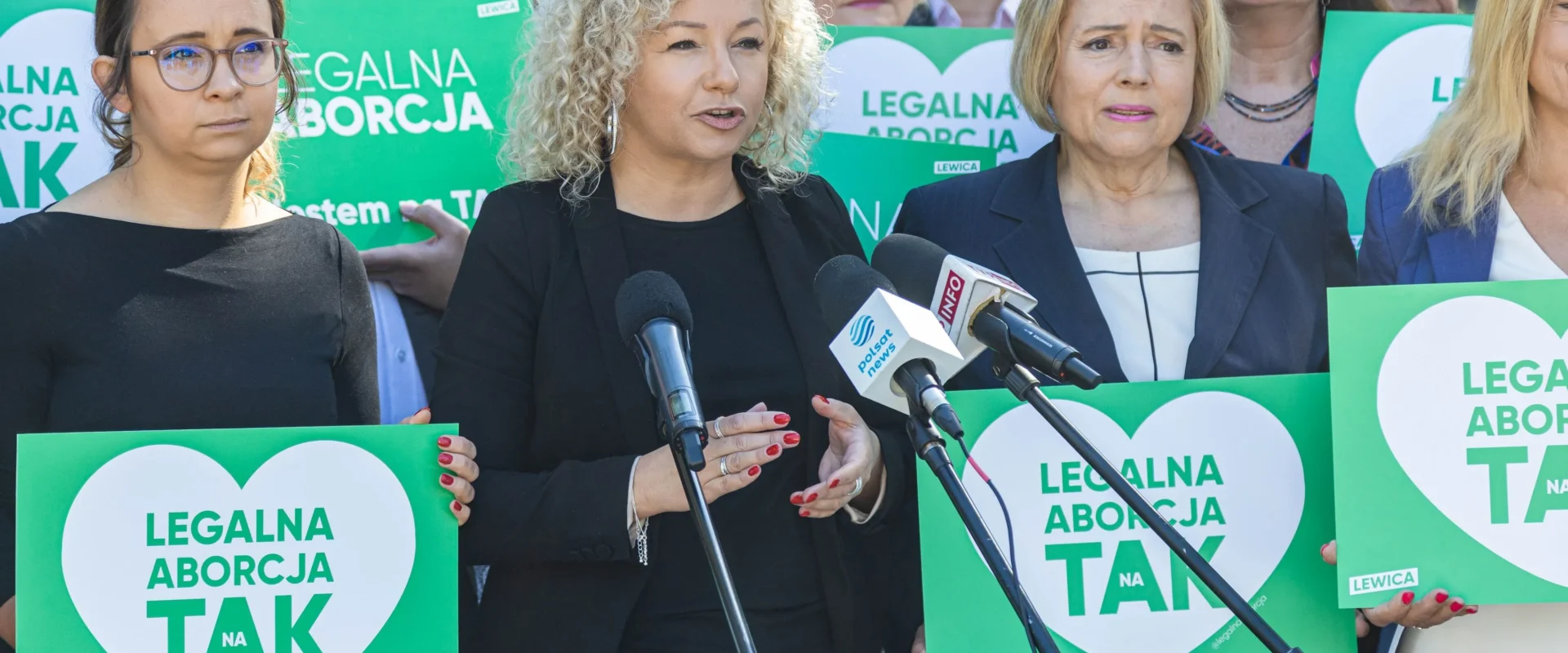 Grupa osób na konferencji prasowej stoi przy mikrofonach i trzyma zielone plakaty z napisem „LEGALNA ABORCJA TAK”.