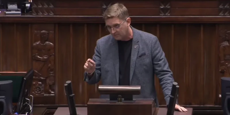 Mężczyzna w szarym żakiecie i czarnej koszulce stoi przy mównicy w sali posiedzeń, gestykulując jedną ręką, z drewnianymi panelami i mikrofonami w tle.