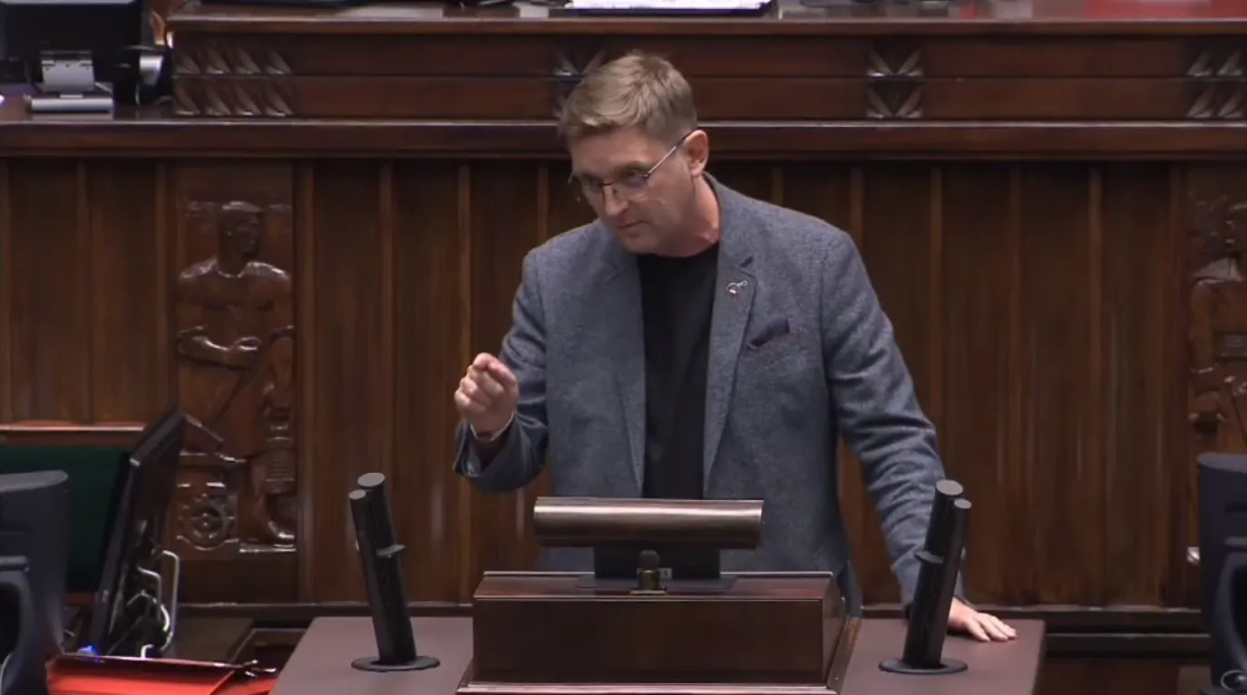 Mężczyzna w szarym żakiecie i czarnej koszulce stoi przy mównicy w sali posiedzeń, gestykulując jedną ręką, z drewnianymi panelami i mikrofonami w tle.