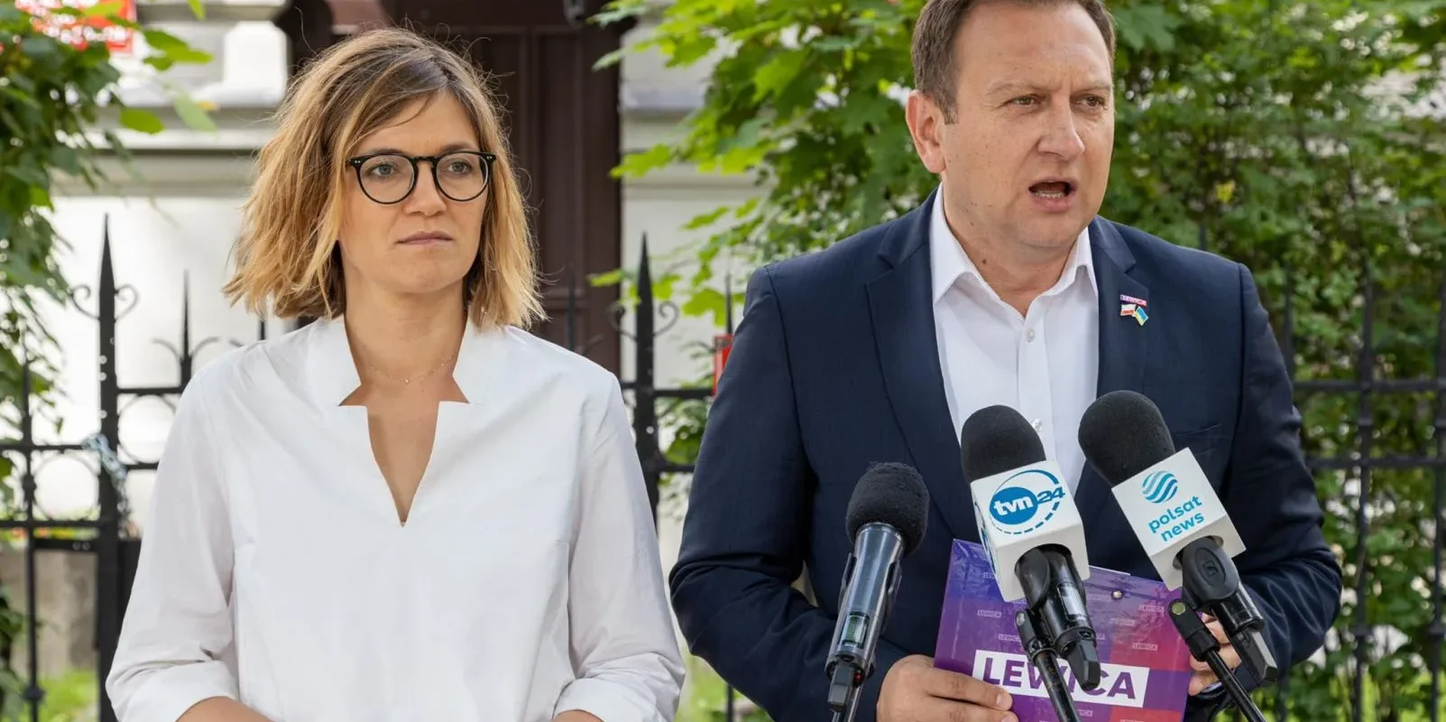 Dwóch polityków stoi na zewnątrz przed mikrofonami podczas konferencji prasowej o inflacji; kobieta w białej koszuli i okularach po lewej, mężczyzna w garniturze po prawej wygłasza oświadczenie, a w tle widać zieleń i ogrodzenie.