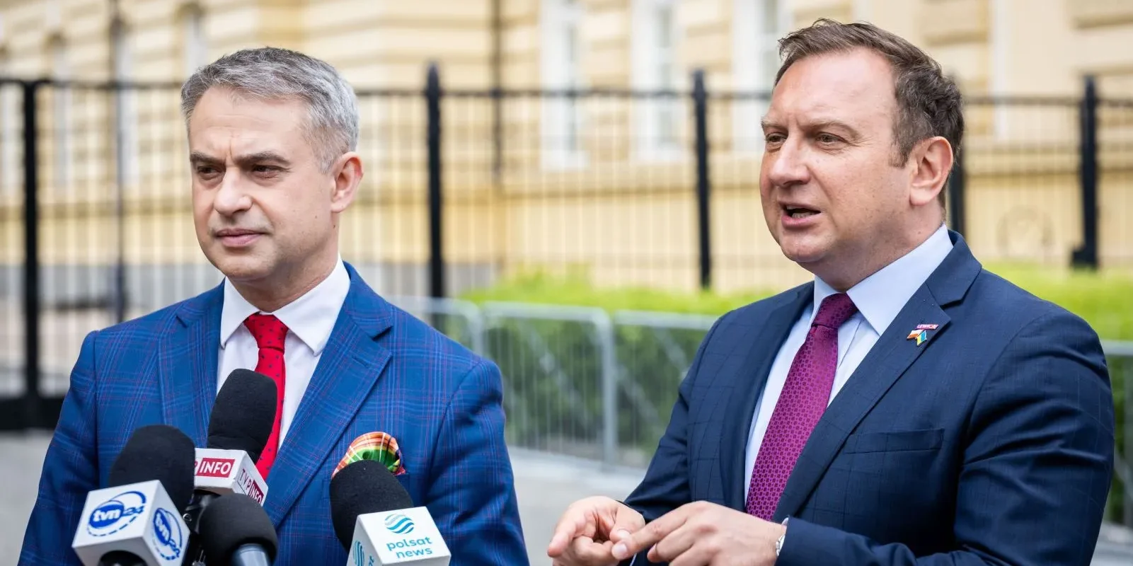 Na zdjęciu dwóch mężczyzn w eleganckich garniturach prowadzi konferencję prasową, stojąc przed budynkiem i mówiąc do zgromadzonych mediów.