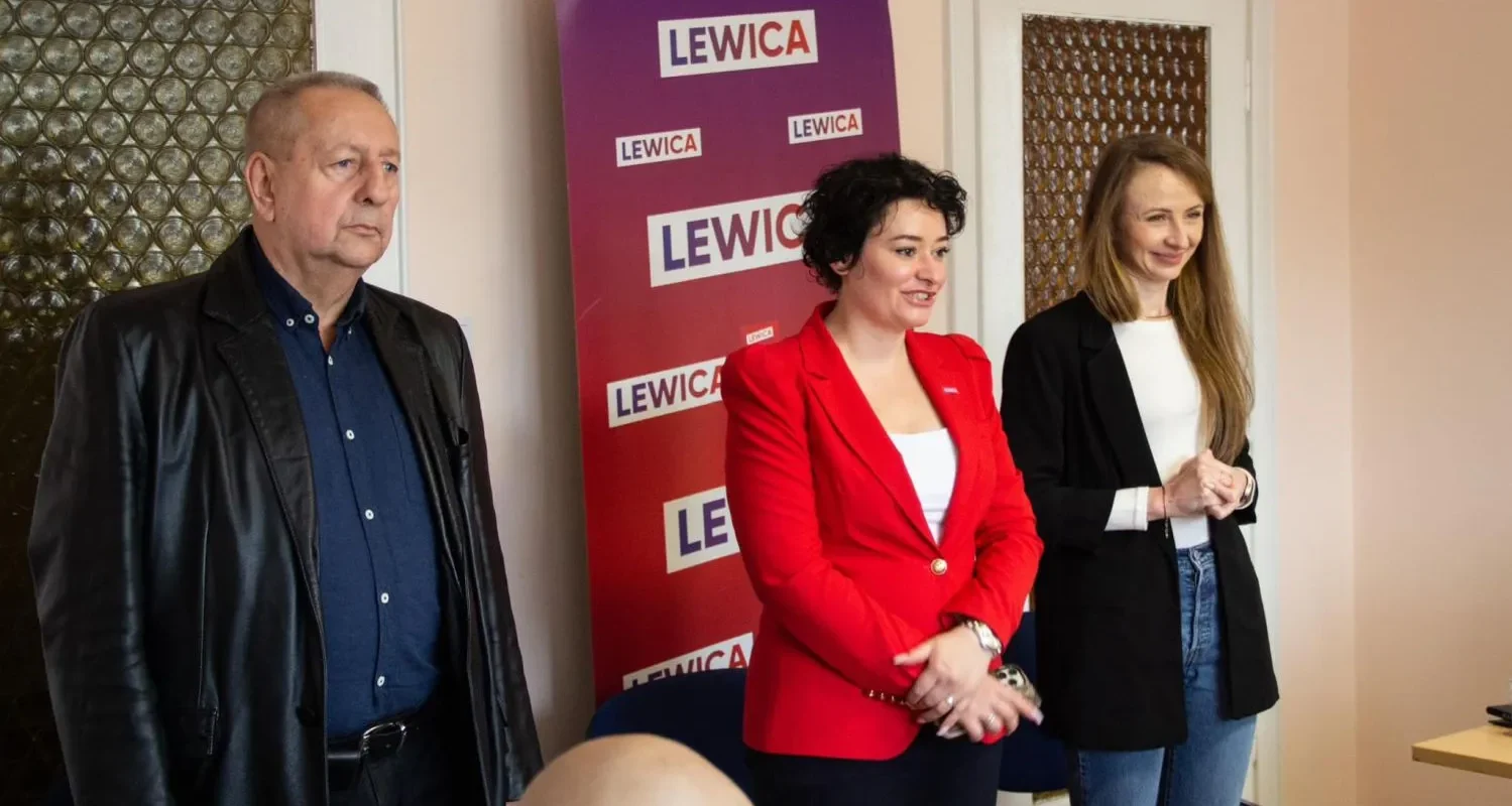 Na zdjęciu przedstawiono troje ludzi stojących przed banerem partii Lewica w biurze, z jedną osobą w czerwonej marynarce na środku.