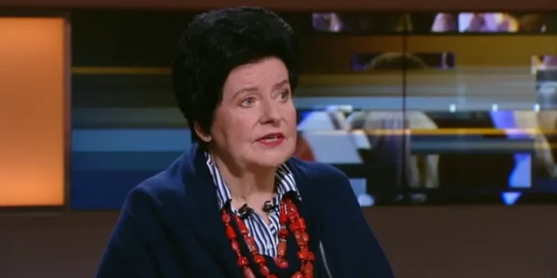 Kobieta z krótkimi czarnymi włosami, ubrana w granatowy żakiet i czerwone korale, siedzi w studiu telewizyjnym.