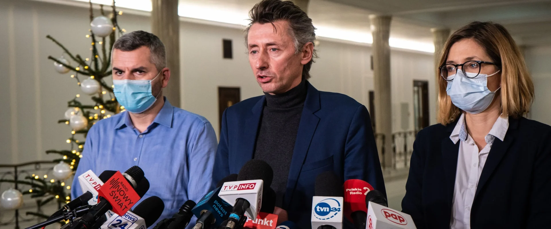 Na zdjęciu widoczna jest trójka osób w maseczkach ochronnych, które prowadzą konferencję prasową w pięknie udekorowanej przestrzeni, w tle widać choinkę.
