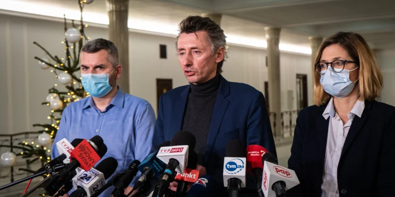 Na zdjęciu widoczna jest trójka osób w maseczkach ochronnych, które prowadzą konferencję prasową w pięknie udekorowanej przestrzeni, w tle widać choinkę.