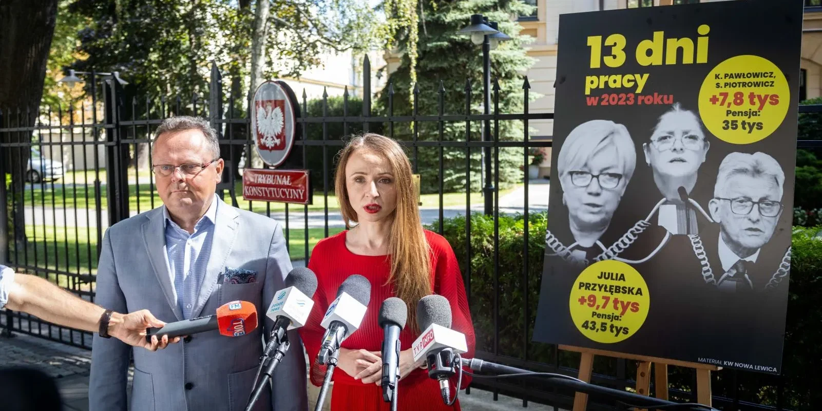 Para osób stojąca przy mikrofonach podczas konferencji prasowej, obok duży plakat z informacjami o pensjach i liczbie dni pracy w 2023 roku.