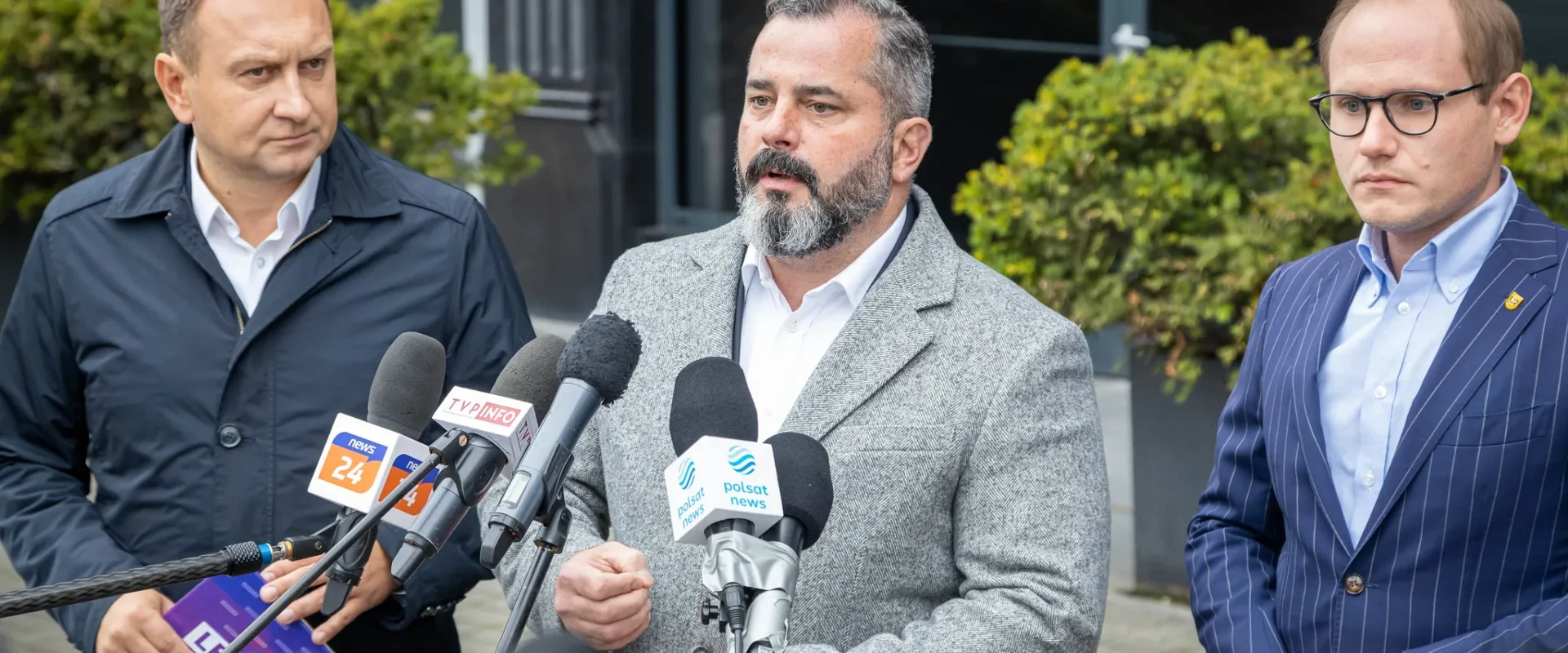 Na zdjęciu trzech mężczyzn stoi przed mikrofonami, prowadząc konferencję prasową na zewnątrz.
