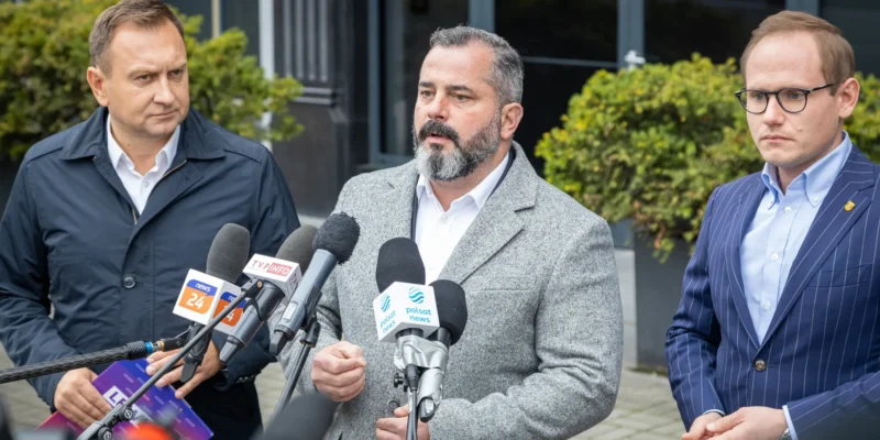 Na zdjęciu trzech mężczyzn stoi przed mikrofonami, prowadząc konferencję prasową na zewnątrz.