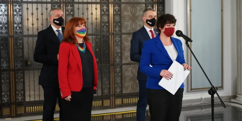 Kobieta w niebieskiej marynarce stoi przy mikrofonie i trzyma kartki, podczas gdy inni w garniturach stoją w tle.