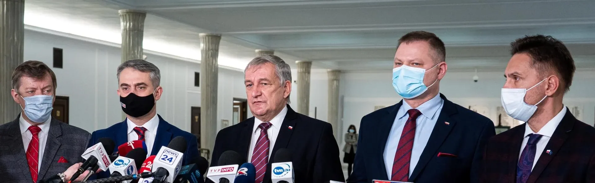 Na zdjęciu widać pięciu mężczyzn w garniturach i maskach, stojących przed mikrofonami w przestronnym wnętrzu budynku.