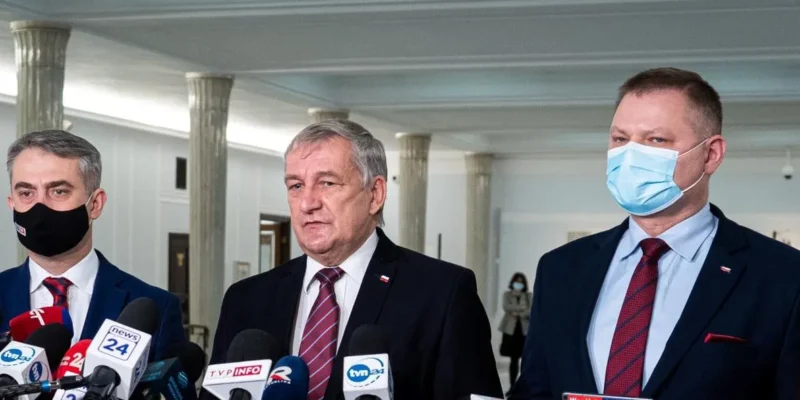 Na zdjęciu widać pięciu mężczyzn w garniturach i maskach, stojących przed mikrofonami w przestronnym wnętrzu budynku.