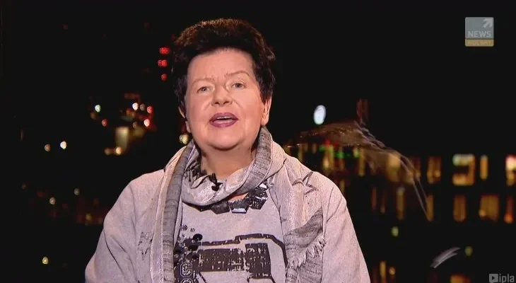 Kobieta mówiąca do kamery podczas wieczornego wywiadu, z przypiętym mikrofonem, na tle rozświetlonego miasta.