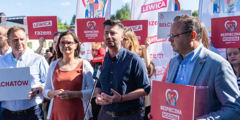 Grupa zwolenników partii Lewica na plenerowym wiecu; mężczyzna w granatowej koszuli przemawia do mikrofonu z czerwonym logo Lewica, a w tle widoczne są plakaty z napisem „Bezpieczna Rodzina” i „Lewica razem”.