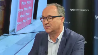 Mężczyzna w marynarce i okularach siedzi w studiu telewizyjnym podczas wywiadu.