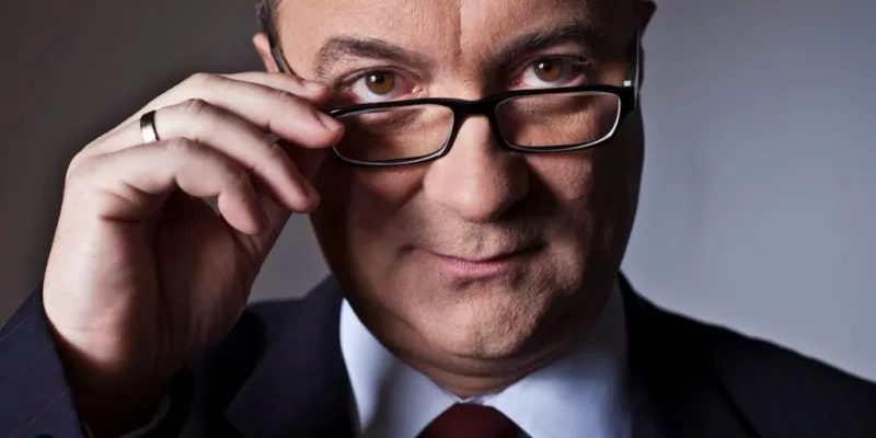 Zbliżenie na mężczyznę w garniturze i krawacie poprawiającego okulary i patrzącego pewnym wzrokiem w obiektyw.