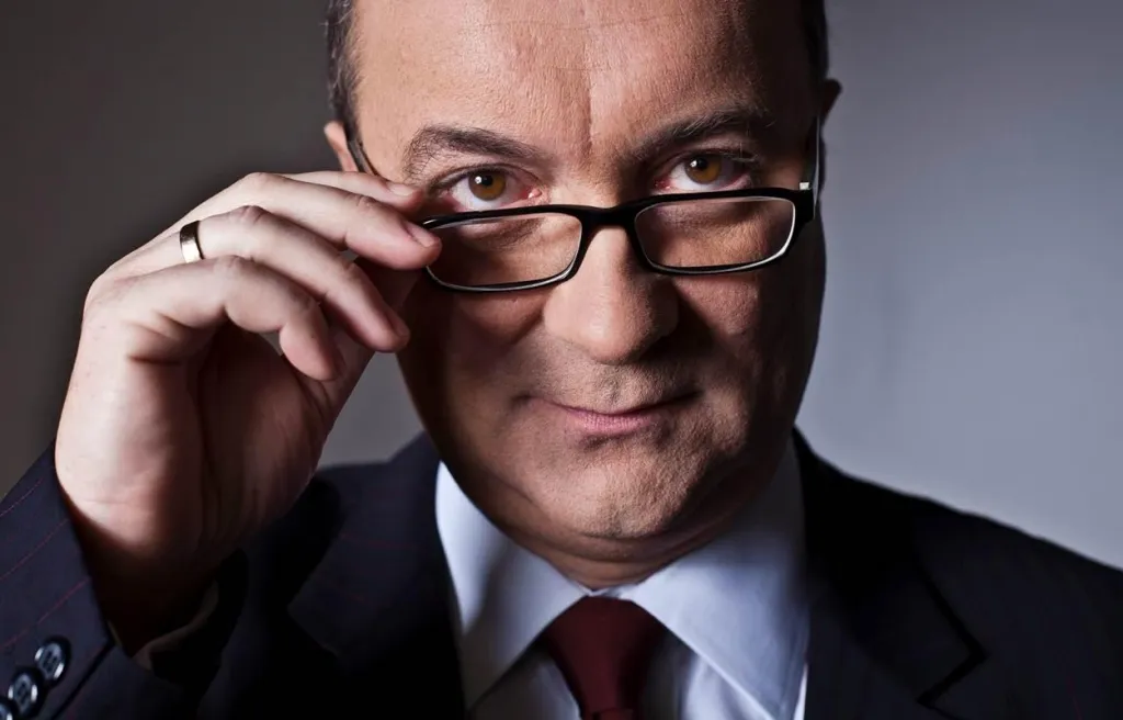 Zbliżenie na mężczyznę w garniturze i krawacie poprawiającego okulary i patrzącego pewnym wzrokiem w obiektyw.