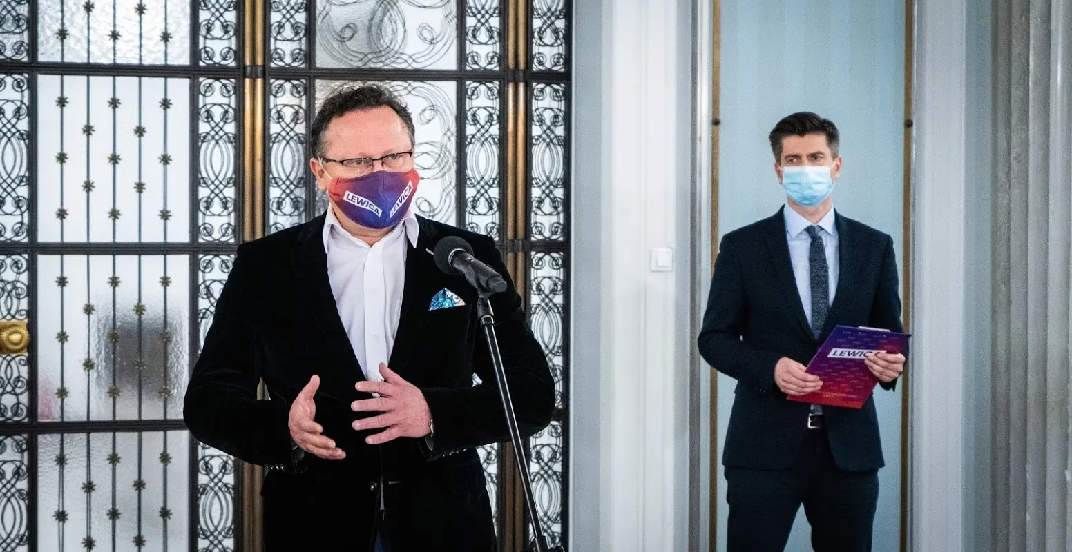 Dwóch mężczyzn w garniturach i maseczkach z napisem LEWICA na konferencji; jeden mówi do mikrofonu, drugi trzyma folder z logo Lewica.