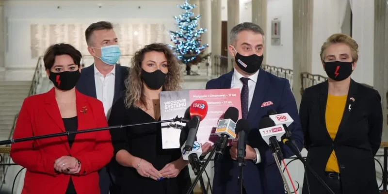 Pięć osób w maseczkach na konferencji prasowej w holu z choinką w tle, stoją przed licznymi mikrofonami, a jedna z nich trzyma plakat.