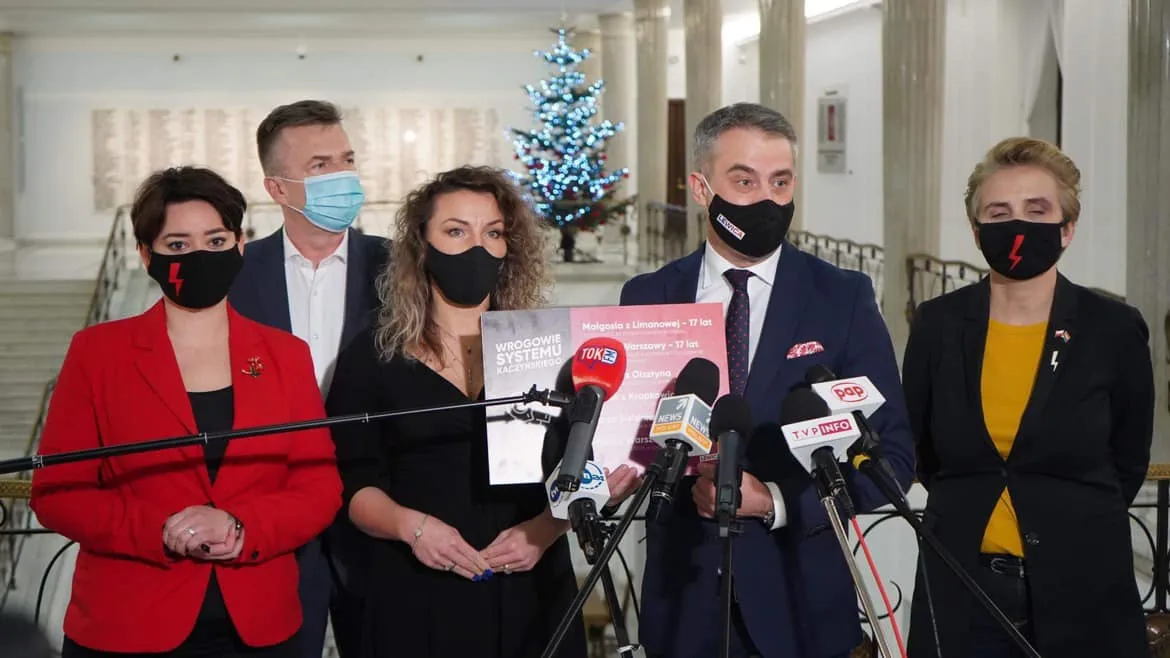 Pięć osób w maseczkach na konferencji prasowej w holu z choinką w tle, stoją przed licznymi mikrofonami, a jedna z nich trzyma plakat.