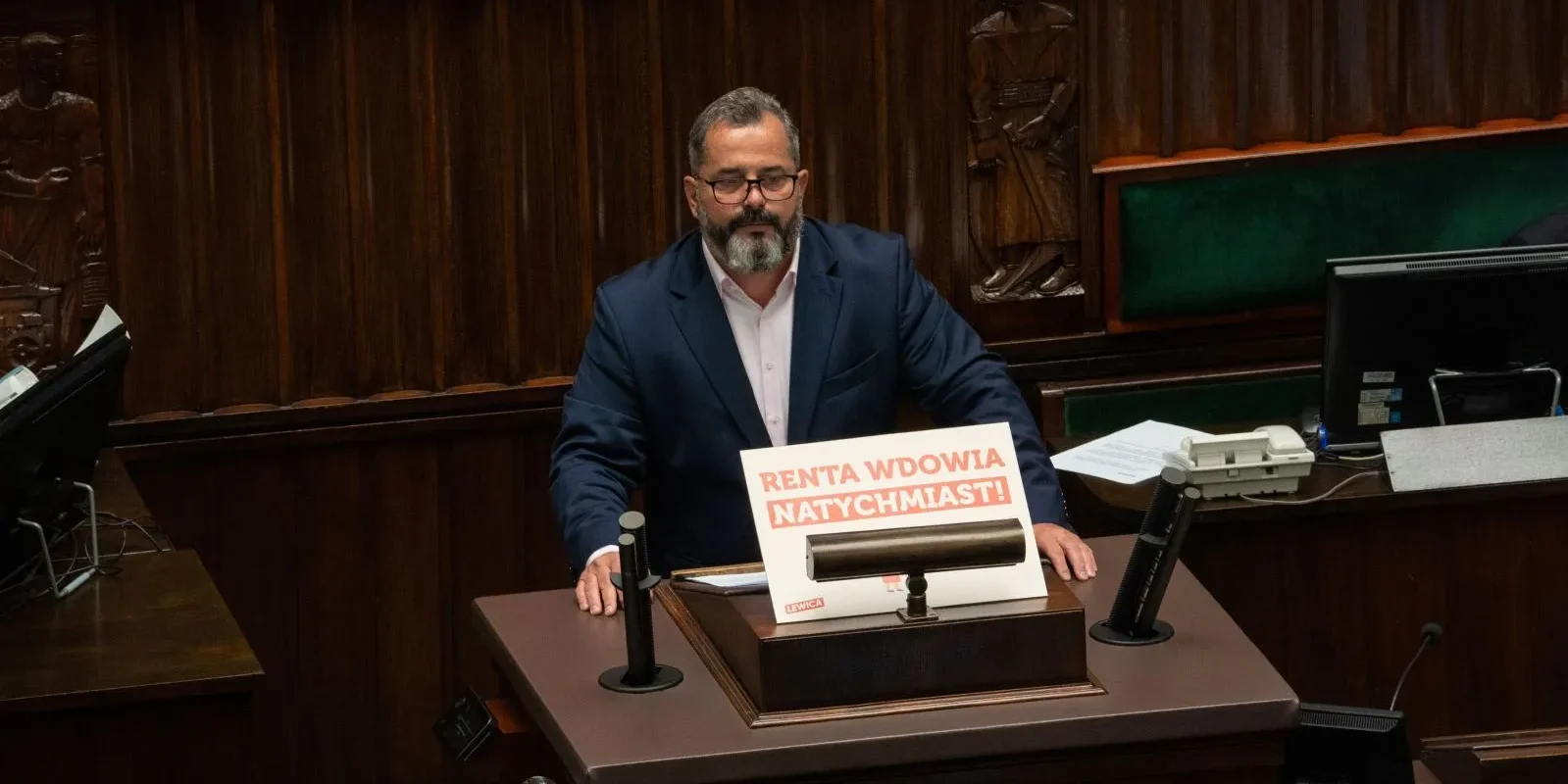 Mężczyzna w garniturze siedzi przy mównicy w drewnianej sali obrad, a na blacie stoi plakat z napisem „Renta wdowia natychmiast!”.