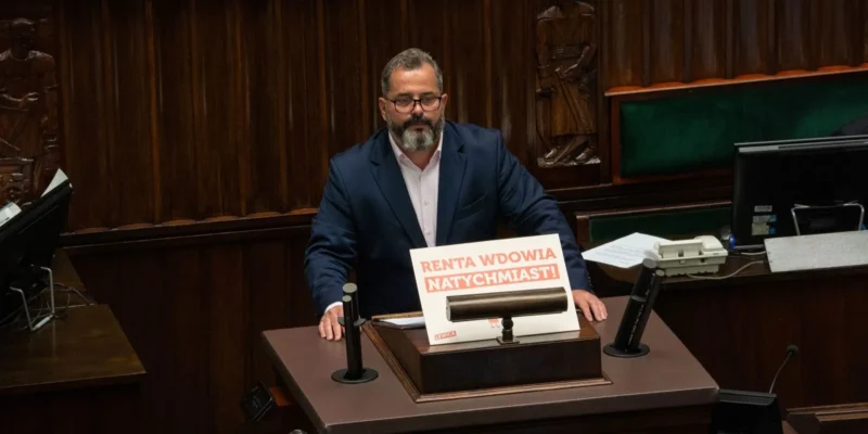 Mężczyzna w garniturze siedzi przy mównicy w drewnianej sali obrad, a na blacie stoi plakat z napisem „Renta wdowia natychmiast!”.