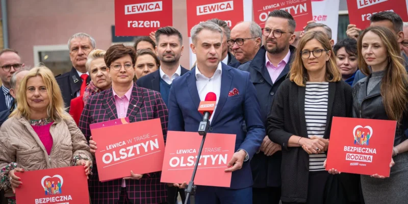Grupa polityków Lewicy podczas konferencji prasowej, na pierwszym planie mężczyzna w granatowym garniturze mówiący do mikrofonu, otoczony innymi uczestnikami i czerwonymi plakatami z hasłami Lewica.