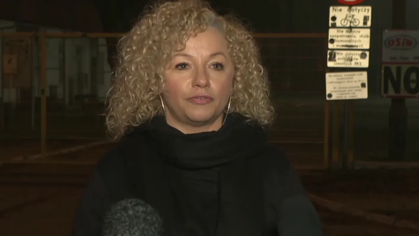 Kobieta o kręconych blond włosach udziela wywiadu na zewnątrz, stojąc przed kamerami i mikrofonami w tle.