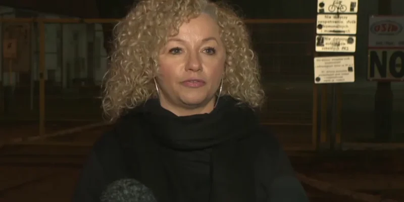 Kobieta o kręconych blond włosach udziela wywiadu na zewnątrz, stojąc przed kamerami i mikrofonami w tle.