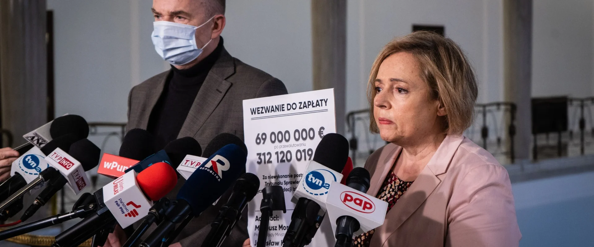 Na zdjęciu widoczni są mężczyzna w maseczce oraz kobieta stojący przed mikrofonami, trzymający plakat z wezwaniem do zapłaty kwoty 69 milionów euro.