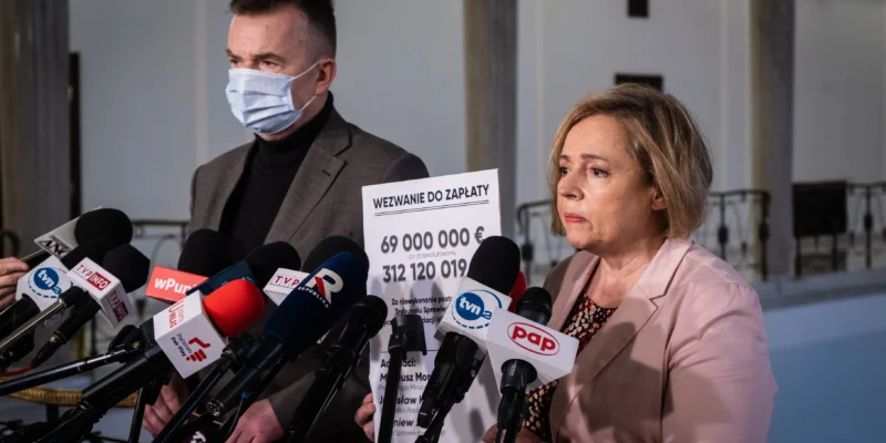 Na zdjęciu widoczni są mężczyzna w maseczce oraz kobieta stojący przed mikrofonami, trzymający plakat z wezwaniem do zapłaty kwoty 69 milionów euro.