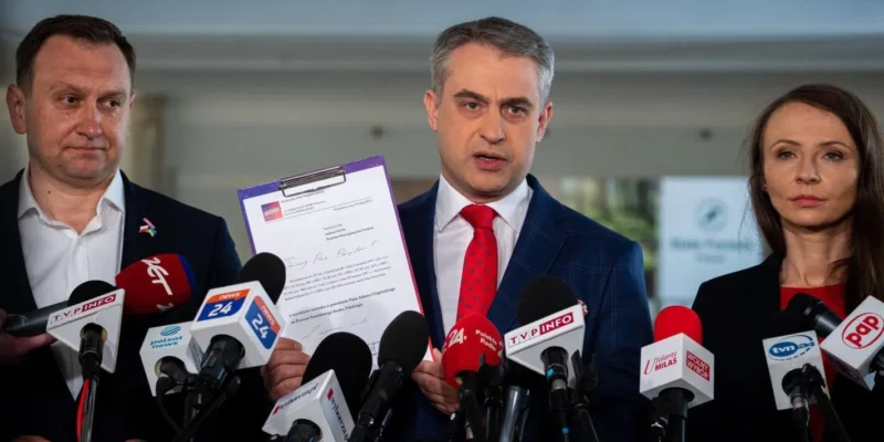 Trzech uczestników konferencji prasowej w garniturach stoi przed licznymi mikrofonami, a centralnie mężczyzna trzyma dokument.