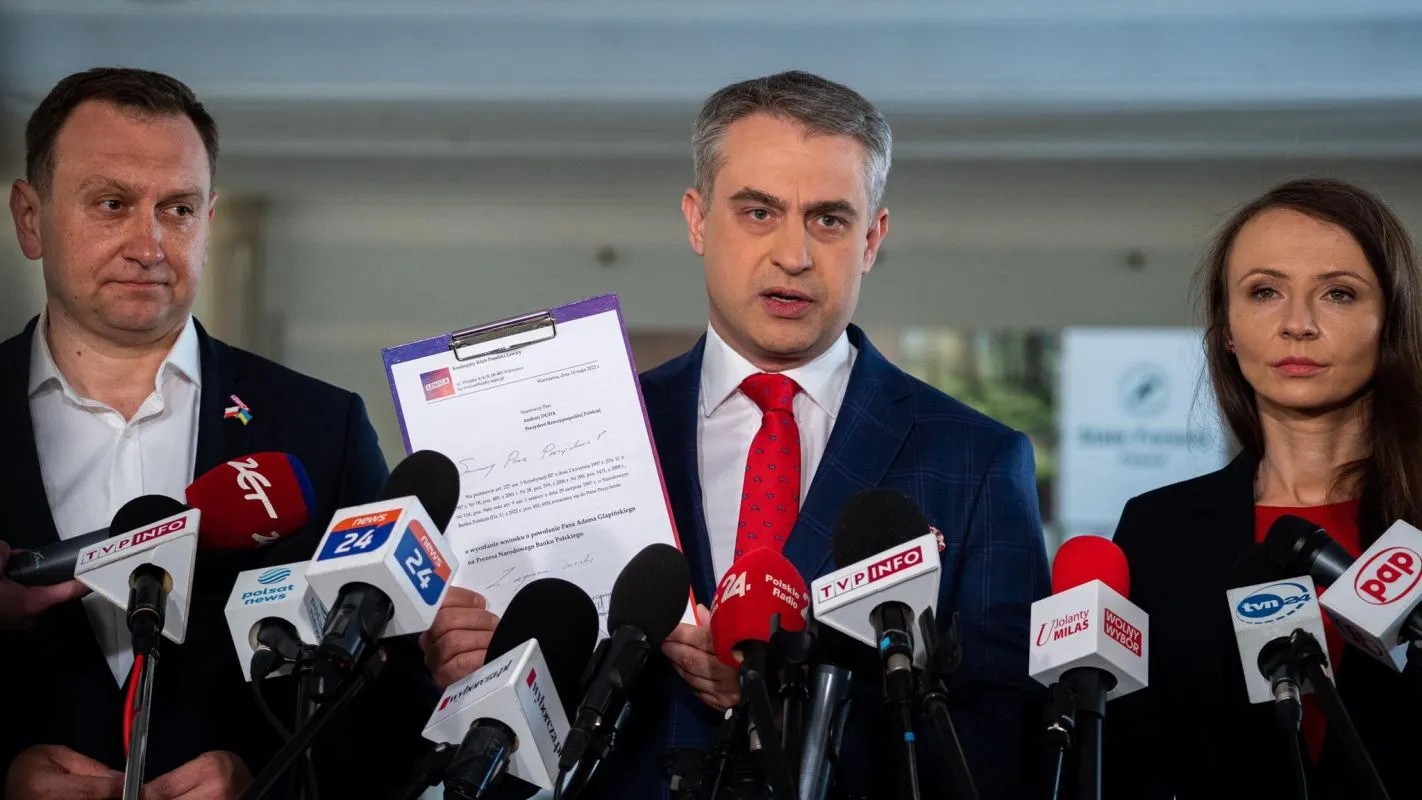 Trzech uczestników konferencji prasowej w garniturach stoi przed licznymi mikrofonami, a centralnie mężczyzna trzyma dokument.