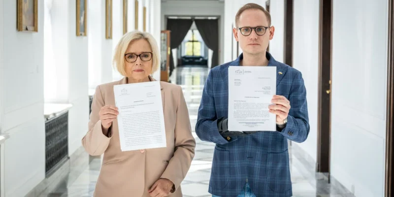 Na zdjęciu w jasnym korytarzu dwóch ludzi trzyma dokumenty i patrzy w stronę obiektywu.