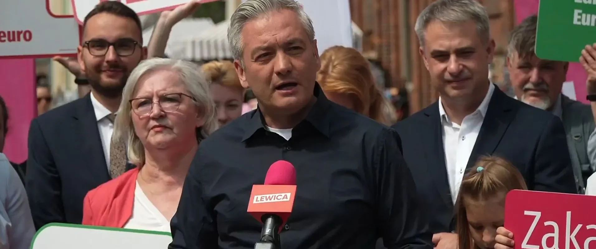 Polityk w czarnej koszuli przemawia do mikrofonu z logiem Lewica podczas zgromadzenia wyborczego, otoczony tłumem i kolorowymi plakatami.